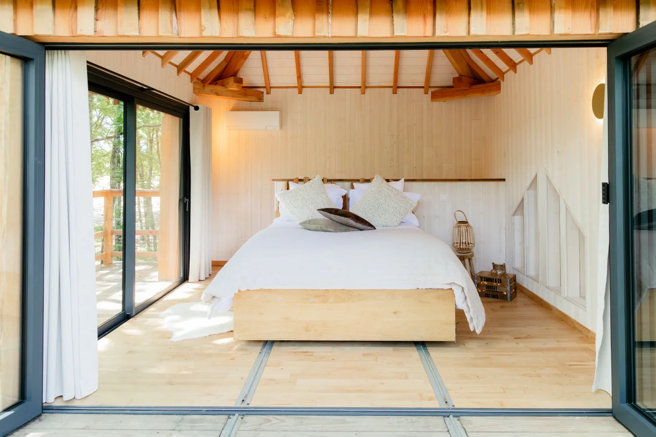 Chambre lumineuse avec lit et grandes fenêtres ouvertes - Cabane dans les arbres Toulouse - Dolce & Cabane